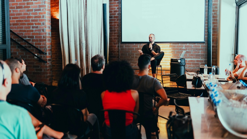 Man sitting on a stage and speaking into a microphone in front of a group of people.